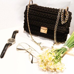 May include: A black crocheted handbag with a gold chain strap and a gold clasp. The bag is sitting on a white surface with a pair of glasses and a bouquet of white flowers.