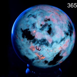 Puede incluir: Una esfera mineral que muestra propiedades fluorescentes bajo luz de 365nm. La superficie presenta un patrón moteado de azul claro brillante, rosa vibrante y parches negros profundos, con un pequeño número 3 escrito a mano en el frente.
