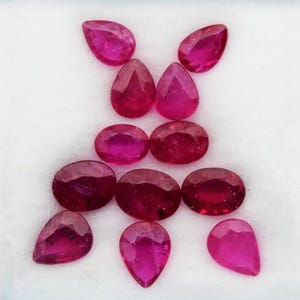 May include: A collection of eleven faceted loose gemstones in a vibrant magenta-red hue. The stones are arranged in a symmetrical pattern, featuring a mix of pear-shaped and oval-cut gems with polished surfaces that reflect light across their facets.