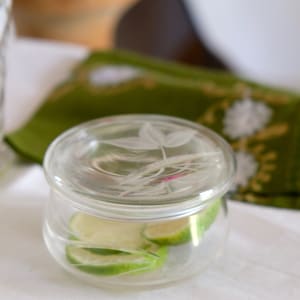 Puede incluir: Un tarro de vidrio transparente con tapa, lleno de rodajas de lima. La tapa tiene un diseño grabado de una hoja.