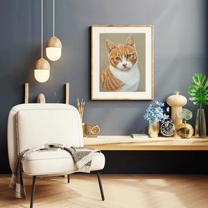 Puede incluir: Una ilustraci&oacute;n enmarcada de un gato jengibre y blanco con ojos verdes, sentado y mirando directamente a la c&aacute;mara. El marco es de madera y tiene un acabado marr&oacute;n claro. La ilustraci&oacute;n est&aacute; sobre un fondo beige.