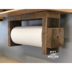 May include: Rustic wooden paper towel holder with a white paper towel roll. The holder is mounted to a wall and has a sign that reads "Pitt and Potter Est. 2018".