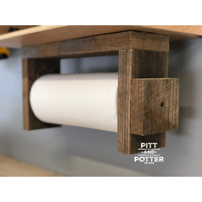 May include: Rustic wooden paper towel holder with a white paper towel roll. The holder is mounted to a wall and has a sign that reads "Pitt and Potter Est. 2018".