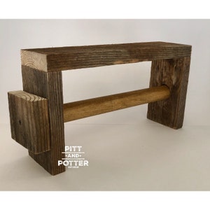 May include: Rustic wooden paper towel holder with a brown wooden dowel. The holder is made of reclaimed wood and has a distressed finish. The holder is labeled "Pitt and Potter Est. 2018".