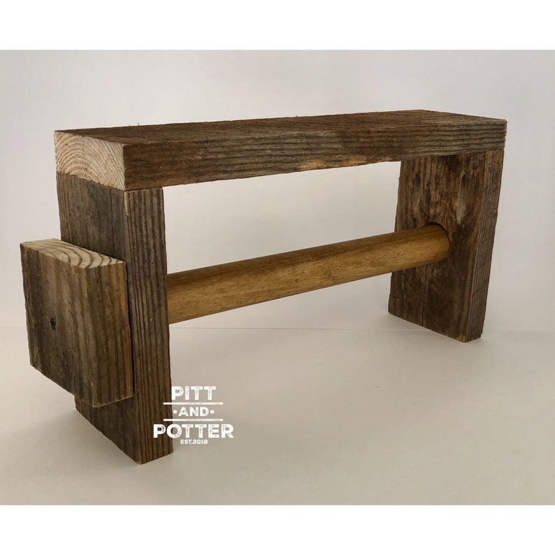 May include: Rustic wooden paper towel holder with a brown wooden dowel. The holder is made of reclaimed wood and has a distressed finish. The holder is labeled "Pitt and Potter Est. 2018".