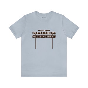 May include: Light blue t-shirt with a brown sign that reads "Welcome to Potter County God's Country".