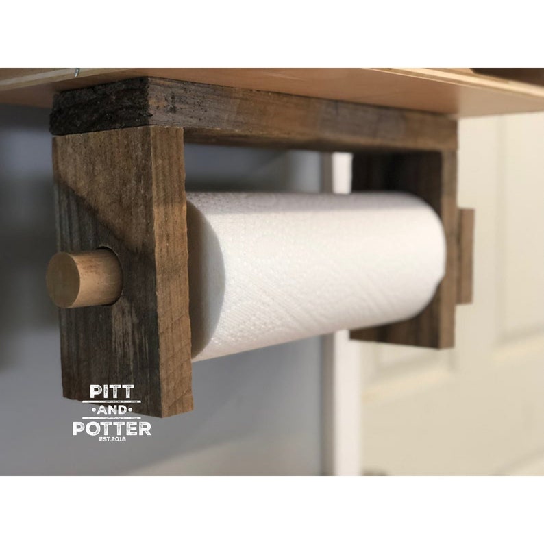May include: Rustic wooden paper towel holder with a white paper towel roll. The holder has a brown wooden dowel and a sign that reads "Pitt and Potter Est. 2018".