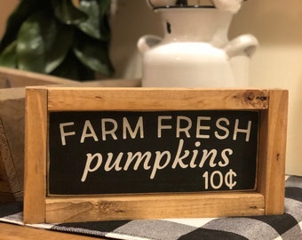 May include: A rectangular wooden sign with the words "FARM FRESH pumpkins 10¢" in white lettering on a black background. The sign is framed in light brown wood and sits on a wooden surface with a black and white checkered runner.