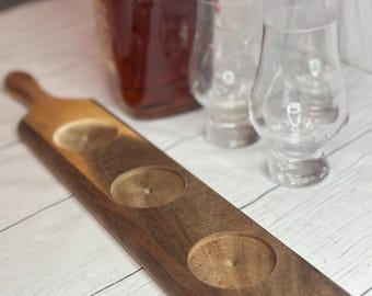 May include: A wooden serving board with two circular cutouts, ideal for holding glasses. The board has a handle and a two-tone finish, with a lighter color on the top and a darker brown on the bottom. Two clear glasses and a bottle are in the background.