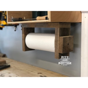 May include: Rustic wooden paper towel holder with a white paper towel roll. The holder is mounted to a wall and has the text "PITT AND POTTER EST.2018" on the bottom.