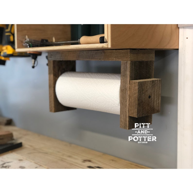 May include: Rustic wooden paper towel holder with a white paper towel roll. The holder is mounted to a wall and has the text "PITT AND POTTER EST.2018" on the bottom.