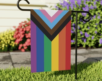 May include: A rainbow pride flag with a brown, light blue, and pink triangle in the top left corner. The flag is attached to a black metal garden flag stand.