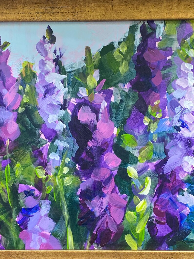 Op de afbeelding: Origineel schilderij van paarse en lavendelkleurige bloemen met groene bladeren. Het kunstwerk is ingelijst in een goudkleurige lijst. De penseelstreken zijn zichtbaar, waardoor een textuureffect ontstaat. De achtergrond is lichtblauw.