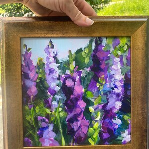 Op de afbeelding: Een ingelijst schilderij van paarse en lavendelkleurige bloemen met groene bladeren. Het kunstwerk wordt gepresenteerd in een goudkleurige lijst. Het schilderij heeft een getextureerde, impressionistische stijl, met zichtbare penseelstreken. De achtergrond is lichtblauw.