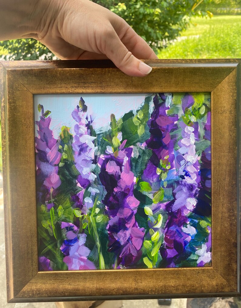 Op de afbeelding: Een ingelijst schilderij van paarse en lavendelkleurige bloemen met groene bladeren. Het kunstwerk wordt gepresenteerd in een goudkleurige lijst. Het schilderij heeft een getextureerde, impressionistische stijl, met zichtbare penseelstreken. De achtergrond is lichtblauw.