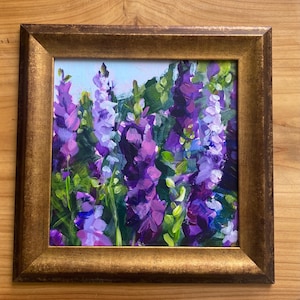 Op de afbeelding: Een ingelijst olieverfschilderij van paarse en violette bloemen met groene bladeren. Het kunstwerk zit in een goudkleurige lijst. De schildering toont een close-up van de bloemen, met een lichtblauwe lucht op de achtergrond.