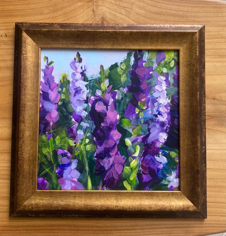 Op de afbeelding: Een ingelijst olieverfschilderij van paarse en violette bloemen met groene bladeren. Het kunstwerk zit in een goudkleurige lijst. De schildering toont een close-up van de bloemen, met een lichtblauwe lucht op de achtergrond.