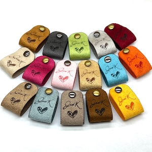 May include: Assortment of 18 leather cord organisers in various colours, each with a different design and a button closure. The designs include hearts, swirls, and abstract patterns.