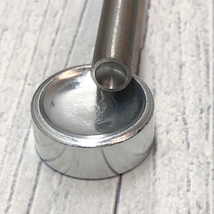 May include: A silver metal punch tool with a round head and a long, cylindrical handle. The tool is resting on a silver metal base.