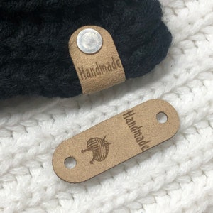 May include: Two brown leather tags with the word "Handmade" and a yarn ball design. The tags are attached to a black knitted hat.