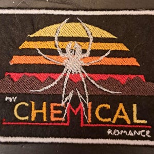 May include: Embroidered patch featuring a spider design over a sunset with the text "CHEMICAL ROMANCE". The patch has a black background with a white border. The spider is white, and the sunset colors are yellow, orange, and red.