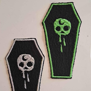 Puede incluir: Dos parches con forma de ataúd. Uno es negro con una calavera blanca y un diseño goteante, contorneado en blanco. El otro es negro con una calavera verde y un diseño goteante, contorneado en verde. Los parches son probablemente para ropa o accesorios.