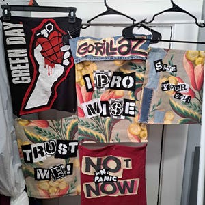 May include: A collection of fabric art pieces, including band-themed designs. One features "GREEN DAY" and a hand holding a grenade. Others display "GORILLAZ", "I PROMISE", "SAVE YOUR SELF", "TRUST ME!", and "NO DON'T PANIC NOW" in bold text.