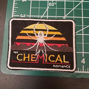 May include: Black embroidered patch featuring a white spider design over a sunset-colored background. The text "MY CHEMICAL ROMANCE" is embroidered in red and yellow. The patch has a white border.