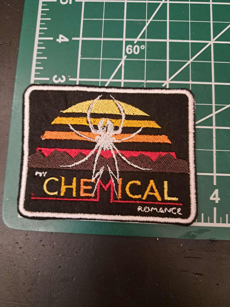 May include: Black embroidered patch featuring a white spider design over a sunset-colored background. The text "MY CHEMICAL ROMANCE" is embroidered in red and yellow. The patch has a white border.