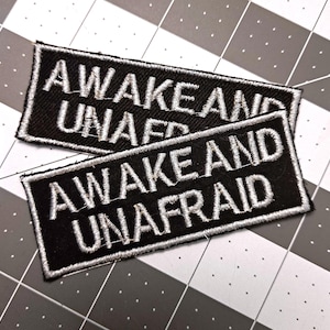 May include: Two black rectangular patches with white embroidered text that reads "AWAKE AND UNAFRAID". The patches have a white border and are placed on a gray and white checkered surface.