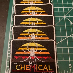 May include: Rectangular black embroidered patches featuring a spider design with a sunset backdrop in yellow, orange, and red. The text "CHEMICAL ROMANCE" is displayed in yellow and red. Patches are stacked.