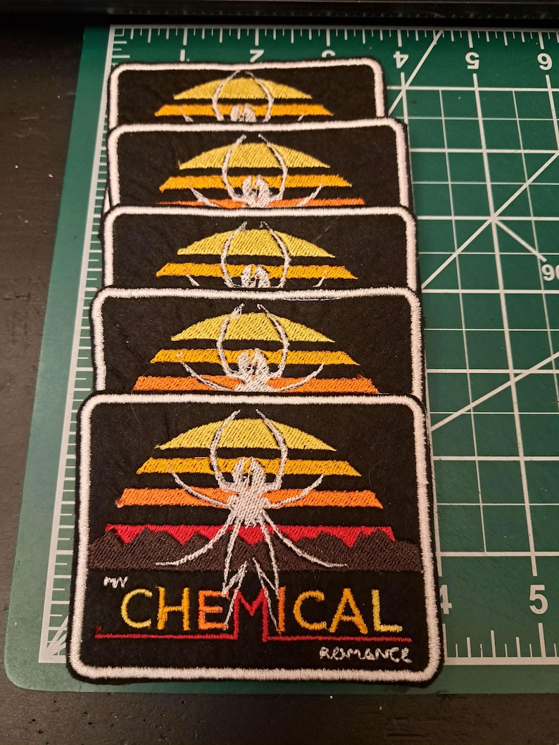May include: Rectangular black embroidered patches featuring a spider design with a sunset backdrop in yellow, orange, and red. The text "CHEMICAL ROMANCE" is displayed in yellow and red. Patches are stacked.