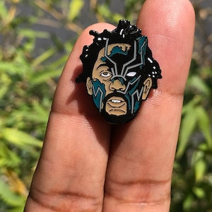 Peut inclure: Épingle en émail noir et bleu sarcelle représentant un portrait stylisé d'une personne avec un design de masque Black Panther. L'épingle est tenue entre deux doigts sur un fond vert flou.