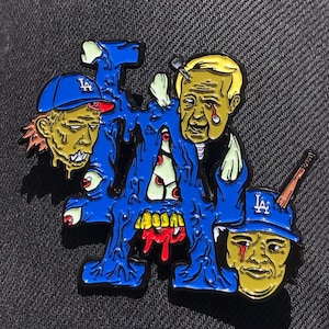 以下が含まれることがあります： 青いエナメルピンは、ゾンビのようなキャラクターが描かれたLAドジャースのロゴの漫画風デザインです。キャラクターは誇張された特徴を持ち、LAドジャースのロゴが入った野球帽をかぶっています。