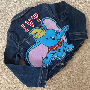May include: A blue denim jacket with a red and white embroidered patch of Dumbo the elephant. The name "IVY" is embroidered on the back of the jacket in red.
