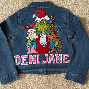 May include: A blue denim jacket with a Grinch, Cindy Lou Who, and Max the Dog Christmas applique. The jacket has the name "Demi Jane" embroidered on the bottom in pink.