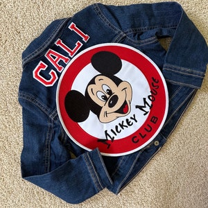 May include: A blue denim jacket with a red and white Mickey Mouse Club patch on the back. The patch has the words "Mickey Mouse Club" written around the edge. The jacket also has the word "CALI" written on the back in red and white letters.