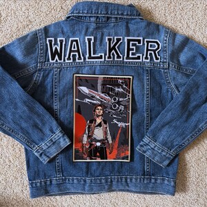 May include: A blue denim jacket with the word "WALKER" in white and black lettering across the upper back. A rectangular patch featuring a Star Wars graphic with a character and spacecraft is on the back.