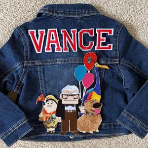 May include: A blue denim jacket with a red and white embroidered name "VANCE" on the back. The jacket has several embroidered patches of characters from the movie Up, including a dog, a bird, and a man with glasses.