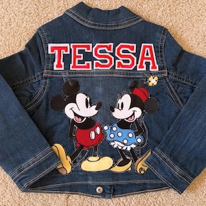 May include: A blue denim jacket featuring Mickey and Minnie Mouse. The back of the jacket has the name "TESSA" in red letters with a white outline. The jacket has a button closure and long sleeves.