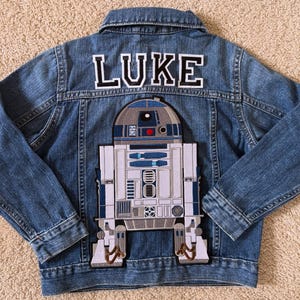 May include: A blue denim jacket with the name "LUKE" in white letters with a black outline. The back of the jacket features an R2-D2 patch in white, blue, gray, and brown.