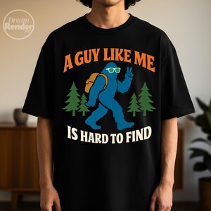 Könnte beinhalten: Schwarzes T-Shirt mit dem Text "A GUY LIKE ME IS HARD TO FIND" in Orange und Weiß. Ein blauer Bigfoot-Charakter mit Sonnenbrille und Rucksack zeigt das Friedenszeichen. Zwei grüne Kiefern umrahmen den Bigfoot.