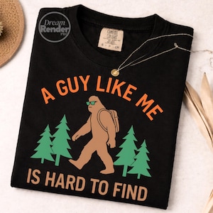 Könnte beinhalten: Schwarzes T-Shirt mit dem orangefarbenen Text "A GUY LIKE ME IS HARD TO FIND". Das Design zeigt einen braunen Bigfoot mit Sonnenbrille und Rucksack, der vor grünen Kiefern läuft. Eine goldene Halskette liegt über dem Shirt.