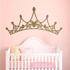 May include: A gold-colored crown wall decal with intricate scrollwork and oval accents is displayed above a white crib with pink bedding and pillows. The wall is painted a soft pink. The decal is a decorative element for a nursery or child's room.