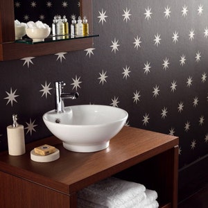 May include: A white ceramic bathroom sink with a chrome tap, a bar of soap, and a dispenser bottle on a dark brown wooden vanity with a shelf. The vanity is in front of a wall with a black and white star pattern wallpaper.