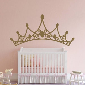 May include: A gold-colored crown wall decal above a white crib with a pink and yellow pillow and a pink bed skirt. The crown has an ornate design with oval accents and a decorative swirl pattern.