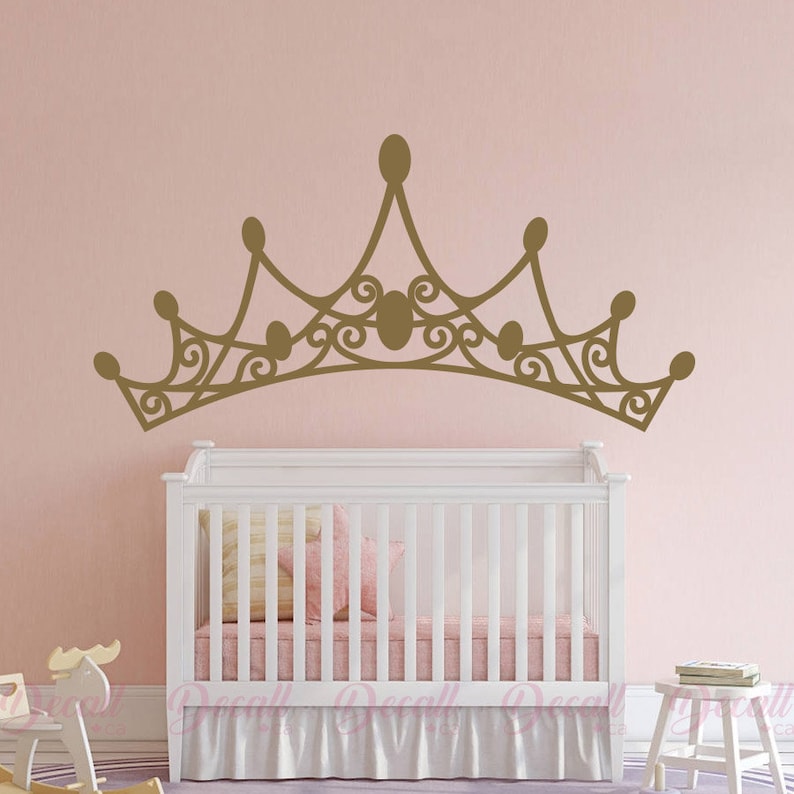 May include: A gold-colored crown wall decal above a white crib with a pink and yellow pillow and a pink bed skirt. The crown has an ornate design with oval accents and a decorative swirl pattern.