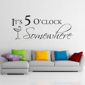 May include: A white wall with a black vinyl decal that reads "It's 5 O'Clock Somewhere" with a martini glass graphic. A grey sectional sofa with colourful throw pillows is in the foreground.