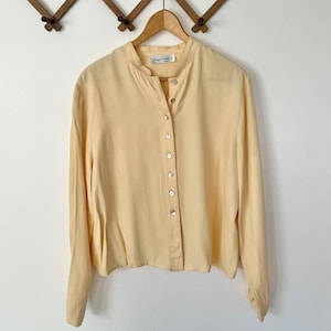 May include: A pale yellow long-sleeved button-up shirt with a mandarin collar. The shirt is hanging on a wooden hanger.