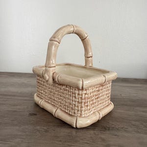 May include: A beige ceramic basket with a bamboo-style handle and trim. The basket has a woven texture on the sides and a rectangular shape. The handle arches over the top, and the interior is a lighter beige color.
